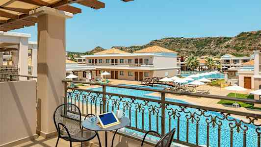 Udsigt fra en balkon til en pool og hotelbygninger med solsenge og parasoller. (AI-genereret tekst)