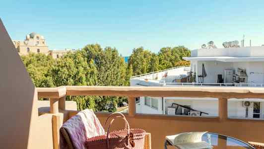 Udsigt fra en balkon mod træer og bygninger. Et bord med en bog og en stol med en taske i forgrunden. (AI-genereret tekst)