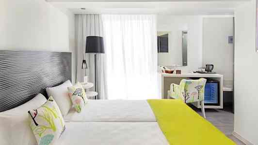 Et lyst hotelværelse med to enkeltsenge, dekorative puder og et limegrønt sengetæppe, samt en siddeplads og et skrivebord med spejl. (AI-genereret tekst)