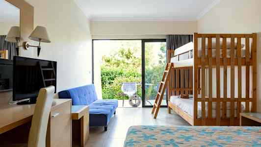 Et hotelværelse med køjeseng, blå sofa, skrivebord, tv og altan med udsigt til grønt. (AI-genereret tekst)