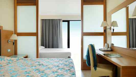 Hotelværelse med dobbeltseng, skrivebord og udsigt gennem et glasvindue. (AI-genereret tekst)