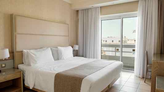 Et hotelværelse med en dobbeltseng beklædt med hvide sengelinned, natborde og store vinduer med gardiner, der lukker lys ind. (AI-genereret tekst)