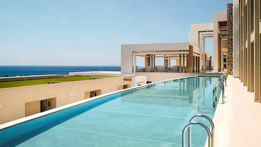 Billede af en pool ved et moderne, lyst bygningskompleks med udsigt over havet. (AI-genereret tekst)