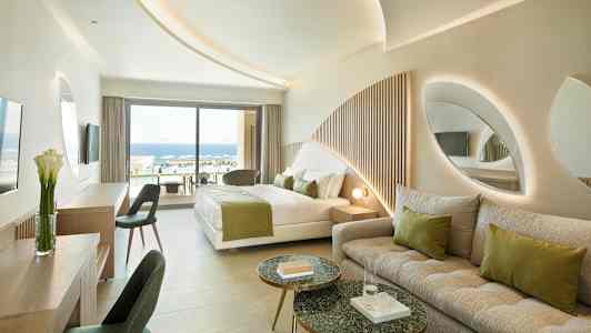 Et moderne hotelværelse med en seng, sofa og udsigt over en pool og havet udenfor. (AI-genereret tekst)