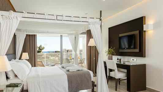 Et elegant hotelværelse med en himmelseng, skrivebord og udsigt til havet fra en altan. (AI-genereret tekst)