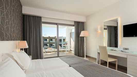 Et hotelværelse med to enkeltsenge, skrivebord, stol og en vinduesudsigt til en balkon med bord og stole. (AI-genereret tekst)