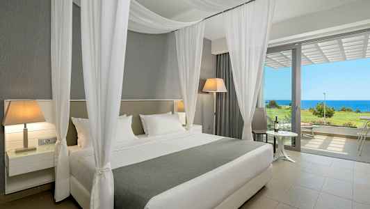 Hotelværelse med en bred seng, hvide gardiner og udsigt til havet gennem store vinduer. (AI-genereret tekst)