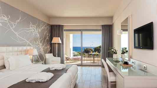 Et hotelværelse med to enkeltsenge, skrivebord, siddeområde og vindue mod en altan med havudsigt. (AI-genereret tekst)