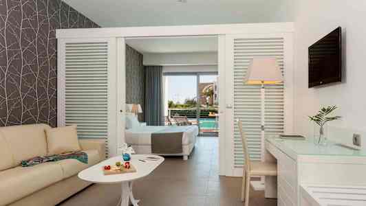 Et hotelværelse med sofa, bord med dekorative genstande, skrivebord og stol. Sengen i baggrunden fører til en terrasse med udsigt over en pool. (AI-genereret tekst)