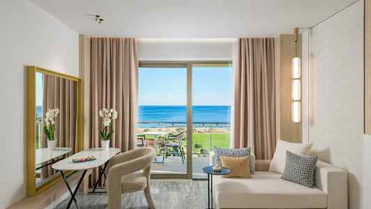 En moderne hotelsuite med sofa, bord, stole og udsigt over havet gennem et stort vindue. (AI-genereret tekst)