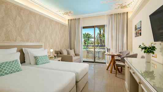 Et hotelværelse med to senge, siddeområde og balkon med udsigt til palmer. (AI-genereret tekst)