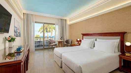 Hotelværelse med to enkeltsenge, siddeområde og altan med udsigt til palmer. (AI-genereret tekst)