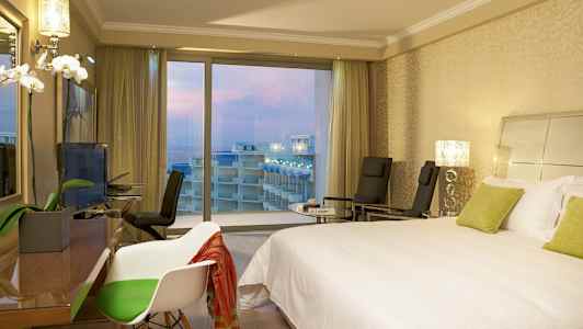 Hotelværelse med en dobbeltseng, stole og udsigt over altaner og havet gennem store vinduer. (AI-genereret tekst)