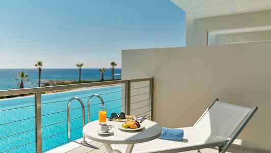 Altan med udsigt over en pool og havet i baggrunden, et bord med morgenmad og en liggestol. (AI-genereret tekst)