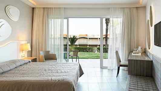 Et hotelværelse med seng, skrivebord, stol og en altandør, der vender mod en have. (AI-genereret tekst)