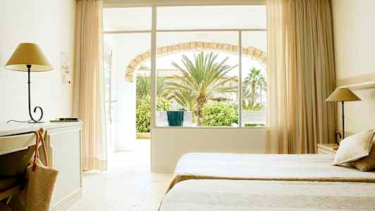 Et hotelværelse med to senge, skrivebord og lampe. Vindue og dør fører til en have med palmer. (AI-genereret tekst)