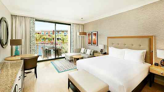 Et hotelværelse med en stor seng, siddeområde og altan med udsigt til en pool og bygninger. (AI-genereret tekst)