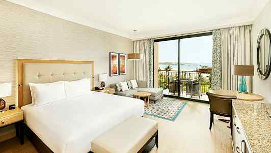 Et hotelværelse med en stor seng, siddeområde, skrivebord og en terrasse med udsigt over havet. (AI-genereret tekst)