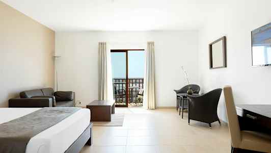 Et hotelværelse med lys indretning. En seng, sofa, bord og stole. En balkon med udsigt ses gennem glasdøre. (AI-genereret tekst)