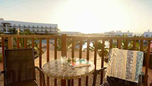 En altan med to stole, et bord og udsigt over en swimmingpool og hotelbygninger. (AI-genereret tekst)