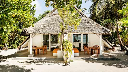 En bungalow med stråtag, omgivet af træer og vegetation, med havudsigt i baggrunden. (AI-genereret tekst)
