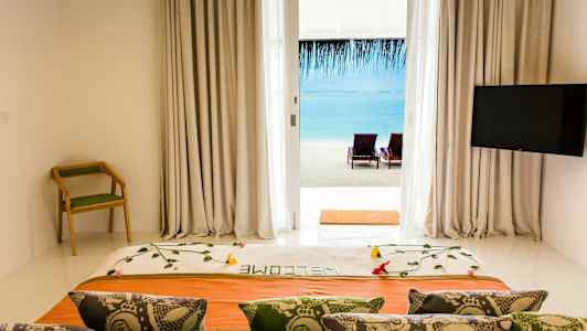 Udsigt fra hotelværelse med dekoreret seng, rygstol og TV mod strand og hav gennem glasdør. (AI-genereret tekst)