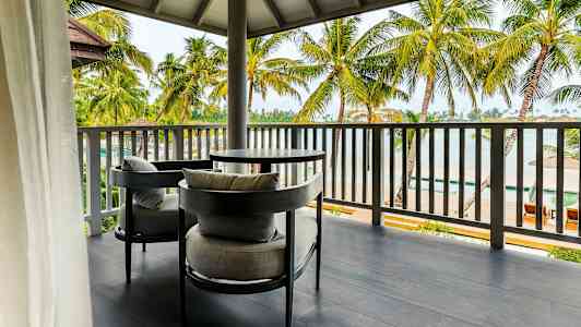 Billede af en udendørsterrasse med to grå siddepladser og et bord. Terrassen har udsigt over palmer og en strand. (AI-genereret tekst)