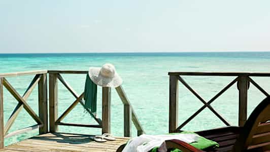 Træterrasse ved havet med en hvid solhat og et grønt klædningsstykke hængende på rækværket. (AI-genereret tekst)