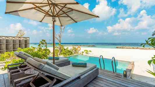 Billede af en solterrasse med parasol og liggestole ved siden af en pool, med hvid sandstrand og hav i baggrunden. (AI-genereret tekst)