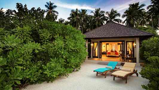 Bungalow foran vegetation med liggestole på sand. (AI-genereret tekst)