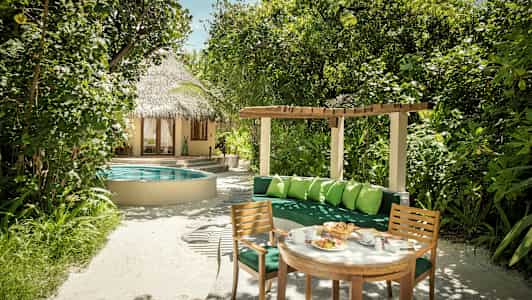 En have med frodige planter, et rundt poolområde, et stråtækket hus og en siddegruppe med bord og stole. (AI-genereret tekst)