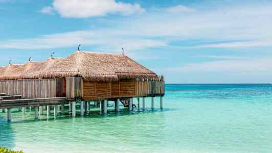 Vandbungalower med stråtag på stolper over turkis vand og klar himmel. (AI-genereret tekst)