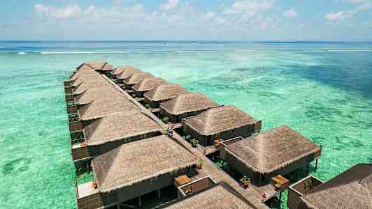 Billedet viser en række vandbungalower med stråtag, bygget på pæle over en turkis lagune. (AI-genereret tekst)