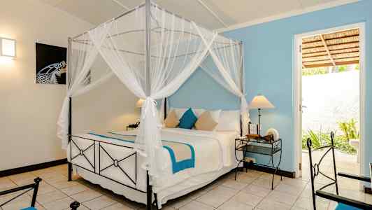 Et soveværelse med en himmelseng med hvide og blå tekstiler, to natborde med lamper og en dør, der fører til et udendørs område. (AI-genereret tekst)