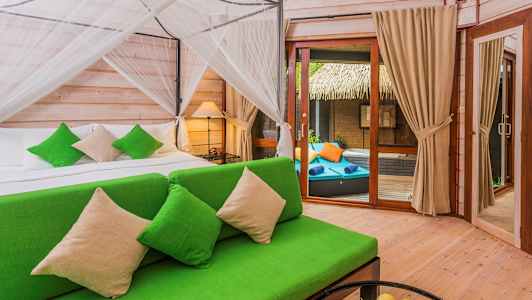 Et soveværelse med en grøn sofa og en seng med grønne pyntepuder. Til højre er en glasdør, der fører til en terrasse med liggestole. (AI-genereret tekst)