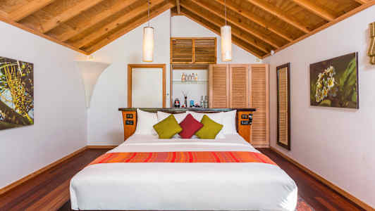 Et soveværelse med træloft, en stor seng med hvidt sengetøj og farverige dekorative puder, samt indretning inklusive lamper og vægdekorationer. (AI-genereret tekst)