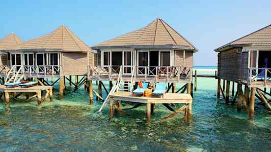 Billedet viser bungalows på pæle over klart vand, med liggestole på en terrasse. (AI-genereret tekst)