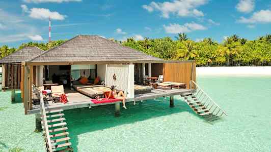 En kvinde sidder på en liggestol ved en luksuriøs bungalow, der er placeret over klart turkis vand, med tropiske træer og en sandstrand i baggrunden. (AI-genereret tekst)