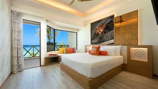 Et moderne hotelværelse med en stor seng, farverige puder, rummelige vinduer og udsigt over havet. (AI-genereret tekst)