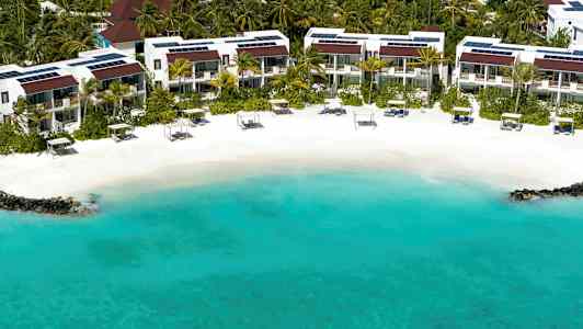 En strand med turkis vand, hvidt sand og liggestole foran et boligområde omgivet af palmer. (AI-genereret tekst)