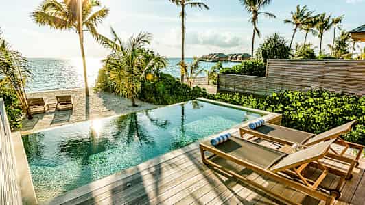 En pool med liggestole på en terrasse ved en sandstrand og havet. (AI-genereret tekst)