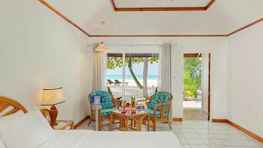 Et hotelværelse med seng, to stole og bord viser udsigt til en strand gennem glasdøre. (AI-genereret tekst)