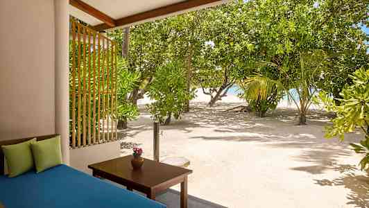 Udsigt fra en terrasse med en blå sofa og et bord med en vase, omgivet af sand og frodige træer nær havet. (AI-genereret tekst)