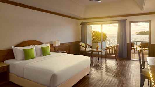 Et hotelværelse med en stor seng, to lænestole og et bord foran en altan med havudsigt. (AI-genereret tekst)