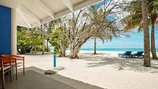 En sandstrand med nogle liggestole, palmer og et blåt hav i baggrunden, set fra en terrasse. (AI-genereret tekst)