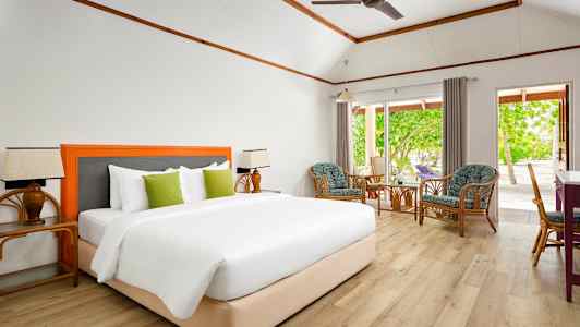 Et værelse med en komfortabel dobbeltseng, siddeområde med stole og et bord, og store vinduer med udsigt til en frodig have. (AI-genereret tekst)