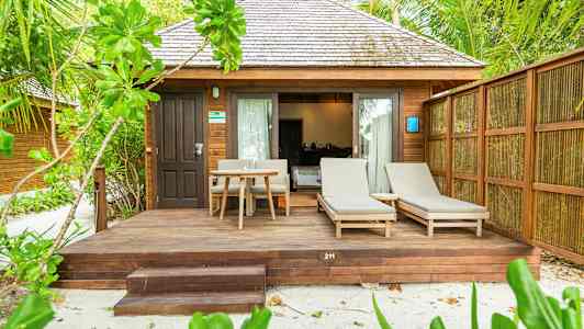 En bungalow med træpaneler, terrasse med liggestole og bord, omgivet af frodighed. (AI-genereret tekst)