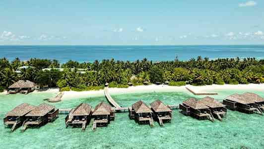 Luksuriøse bungalower på pæle over klart turkis vand og en tropisk strand med palmer. (AI-genereret tekst)