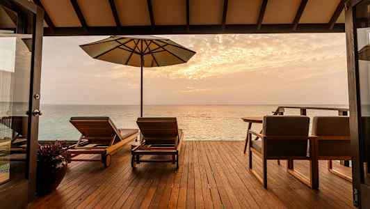 En terrasse med liggestole og parasol, der vender ud mod havet ved solnedgang. (AI-genereret tekst)