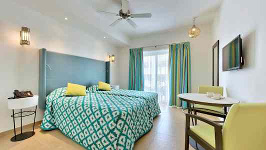 Et hotelværelse med to enkeltsenge, en lille spiseplads og et væg-TV foran vinduer med farverige gardiner. (AI-genereret tekst)